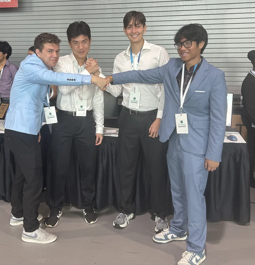 Four SharkMakers students in suits with conference badges, clasping
                hands together at the Synapse Summit exhibition floor.