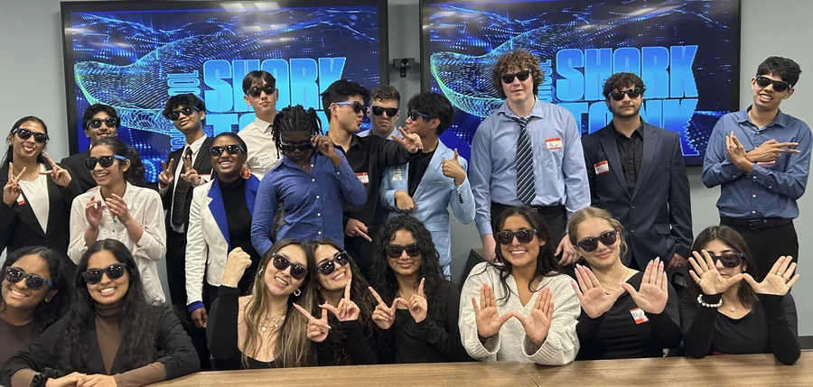 About twenty SharkMakers students in sunglasses, making fin shapes with their hands, under the Shark Tank screens.