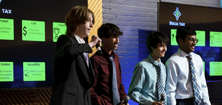 Four students presenting their Ninja Tax pitch on stage, microphones in hand, their slides behind them.